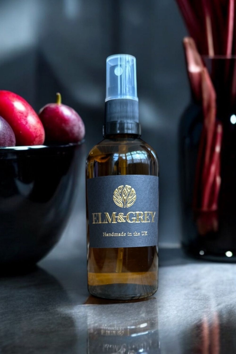 Bottle of Elm & Grey room spray on a reflective surface with apples and a vase in the background