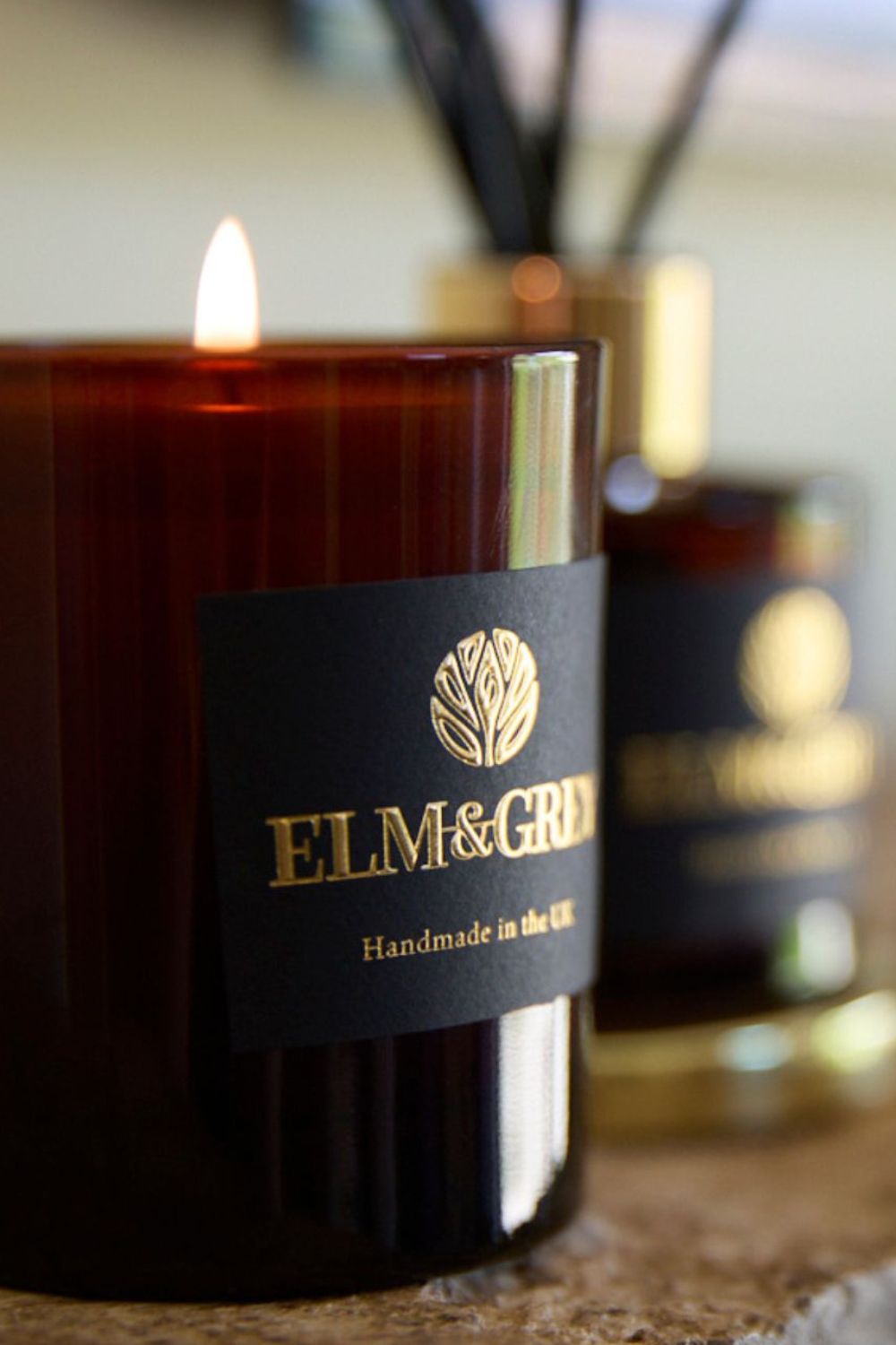 Candle with a visible flame and 'ELM & GREY' label, with an amber diffuser in the background.