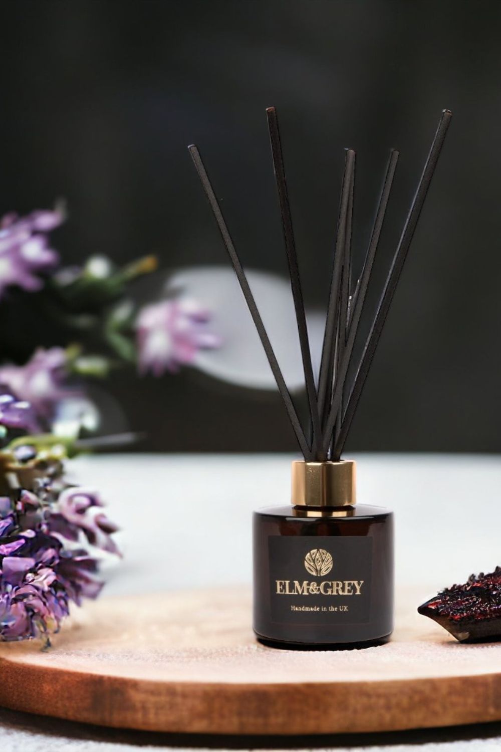Elm&Grey diffuser with reeds on a wooden board with flowers in the background