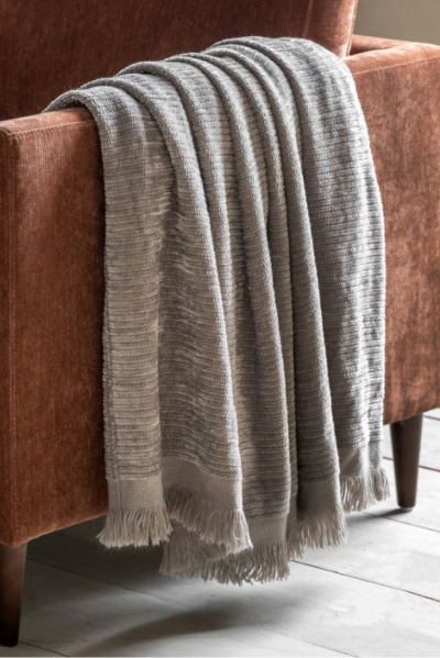 A grey chenille throw blanket draped over the arm of a brown velvet chair, placed on a wooden floor.