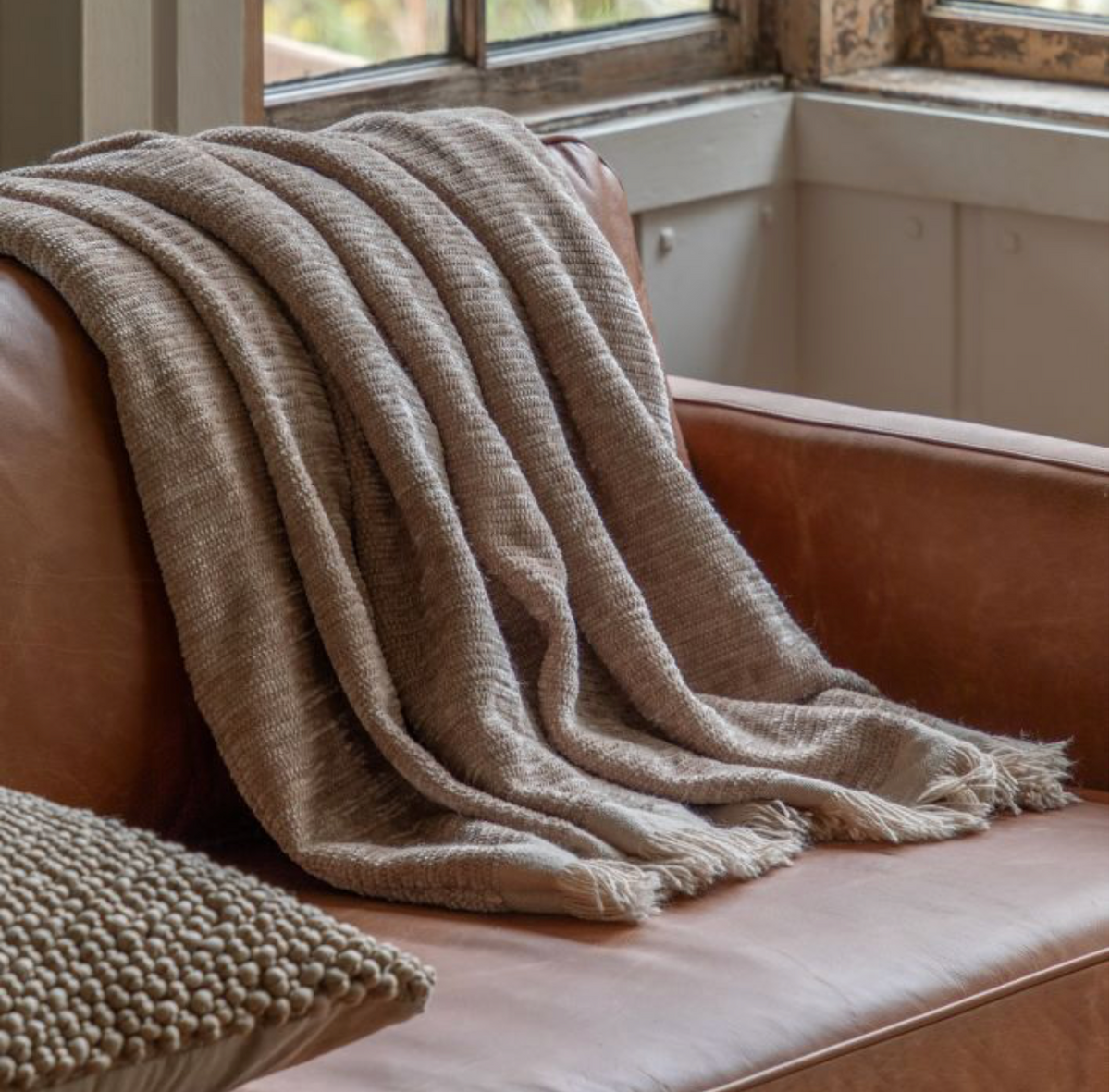 A natural color chenille throw blanket draped over a couch, with a leather couch and a cushion visible.