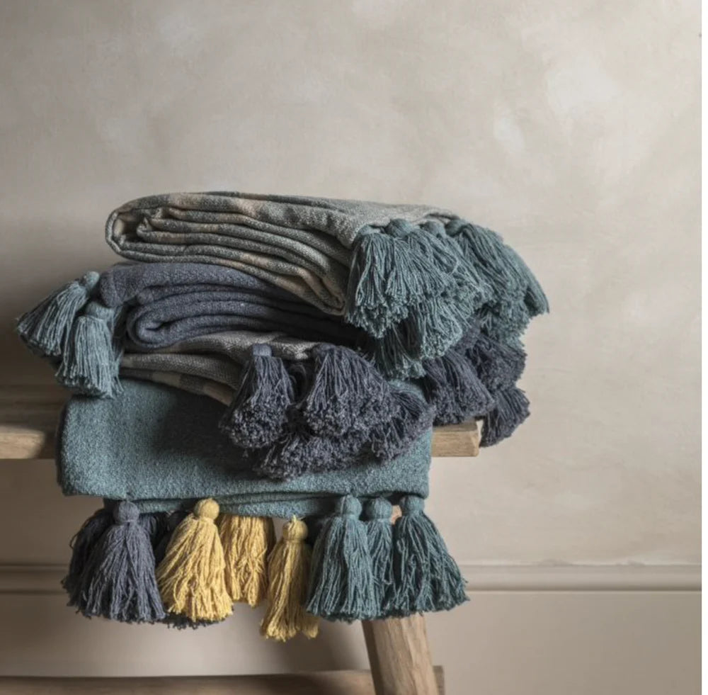 A recycled cotton throw with multicoloured tassels, featuring shades of blue and gray, draped over a wooden stool against a textured wall.