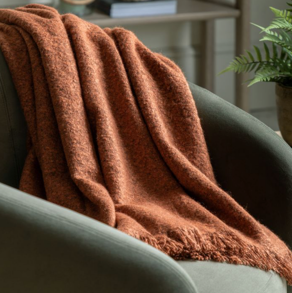 A cosy bouclé-style throw in a warm rust hue draped over a modern chair, with a plant and a bookshelf in the background.
