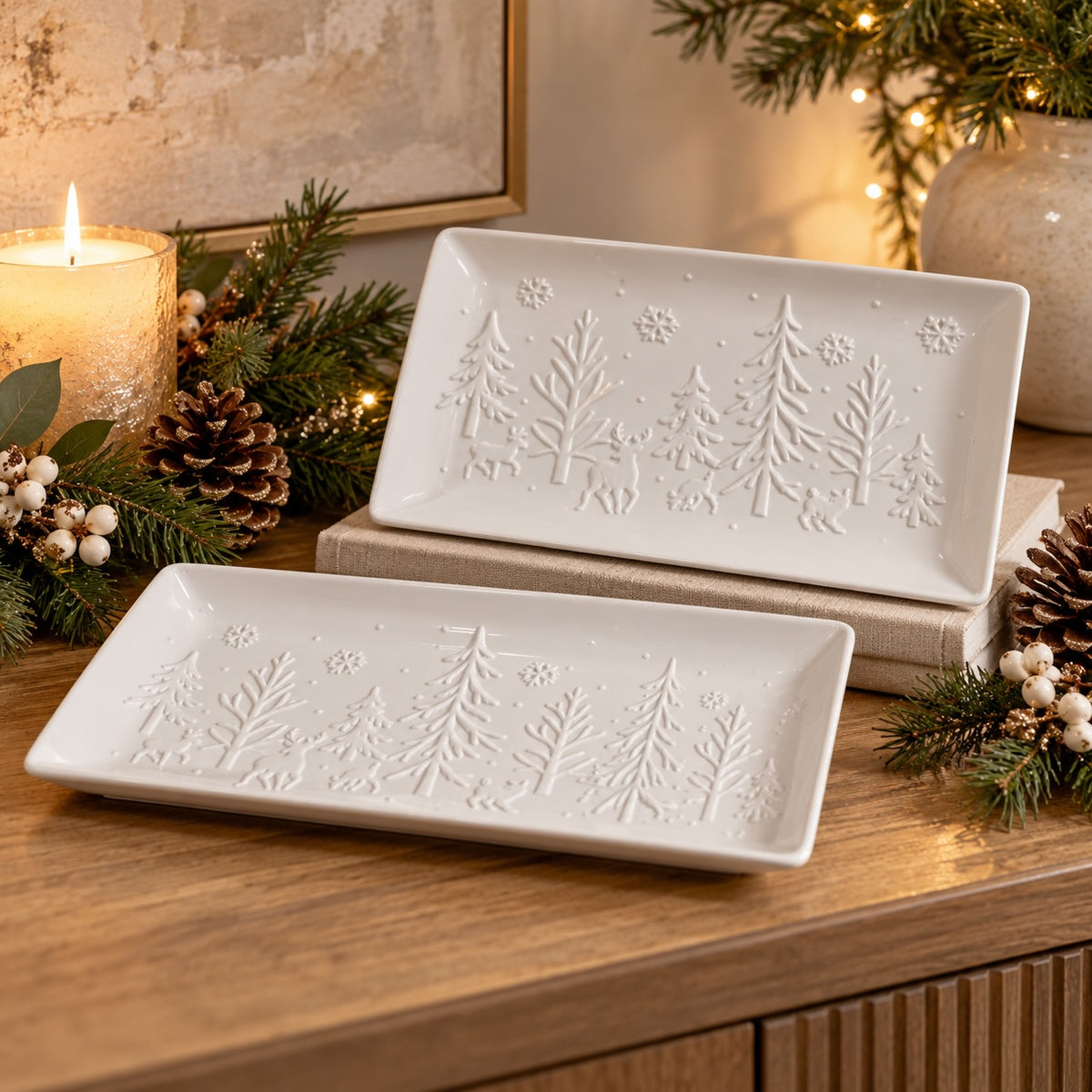 Two white rectangular ceramic serving platters with embossed winter tree designs, styled on a wooden table with candles, pine branches, and warm Christmas lights in the background.