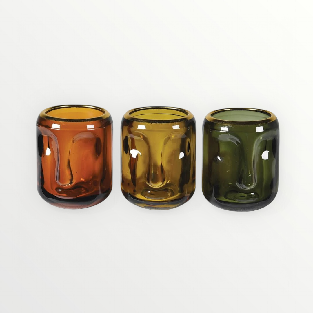 A set of three small glass candle holders in amber, green, and golden yellow colors, with a face design.