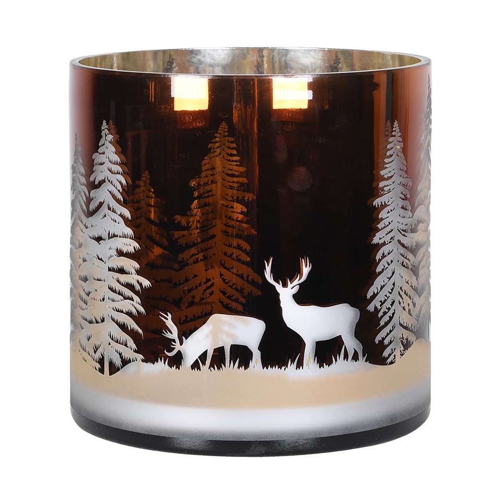 A glass candle holder featuring a winter forest design with white silhouettes of pine trees, a deer, and a stag, shown lit against a white background.