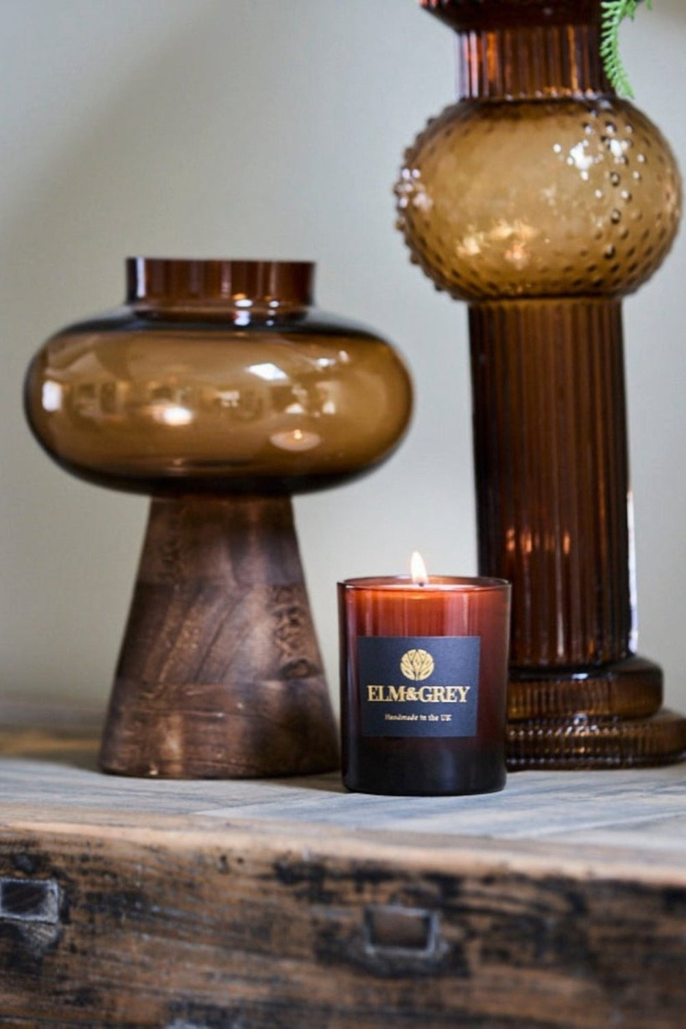 Candle with a 'Ellm&Grey' label between two brown glass lanterns on a wooden surface.