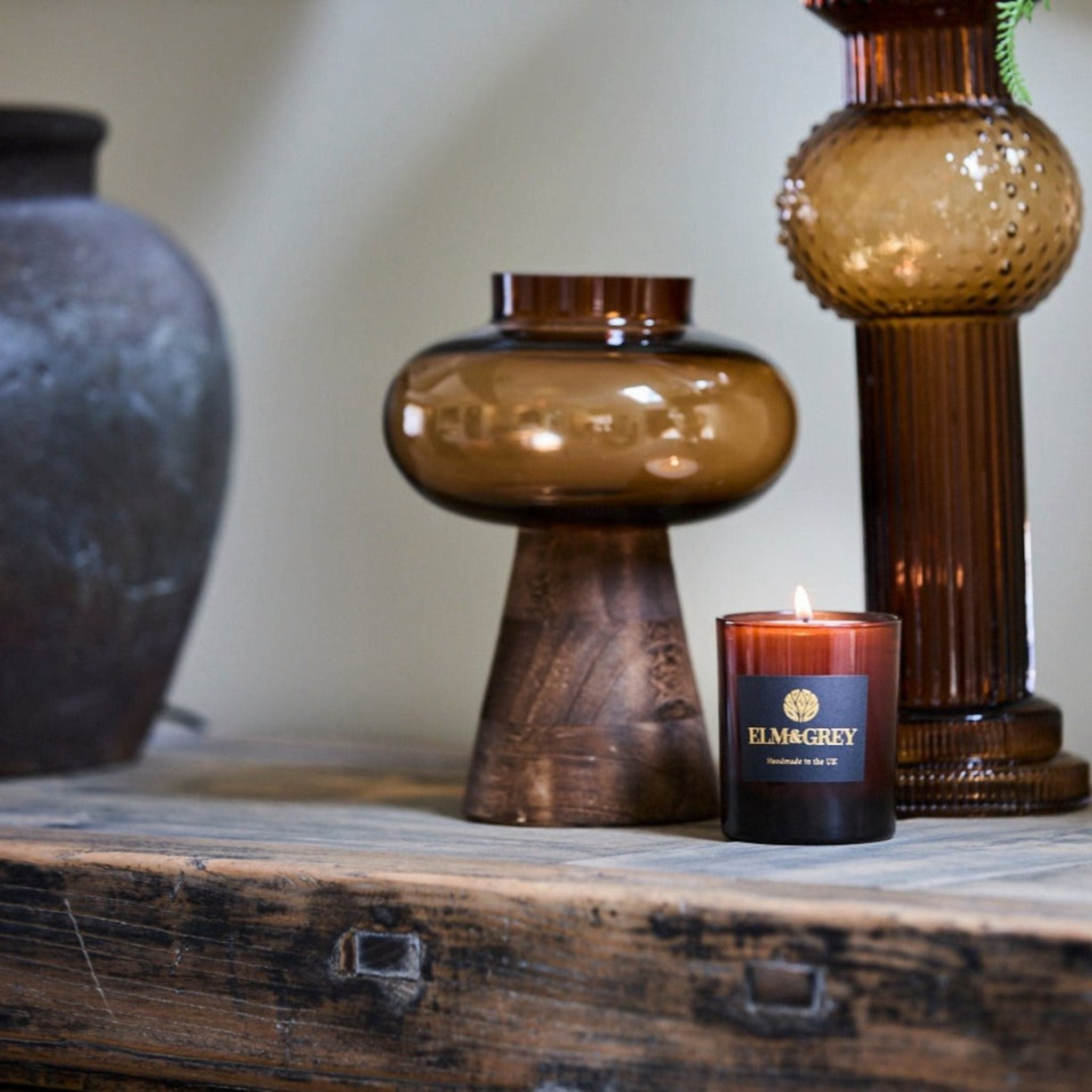 Candle with 'Ellm&grey' label on a wooden surface with decorative vases.