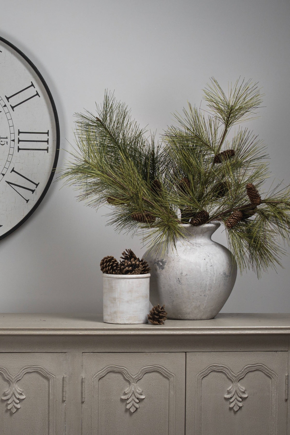 Decorative setting with a vase of greenery, small pot, and clock on a gray surface.