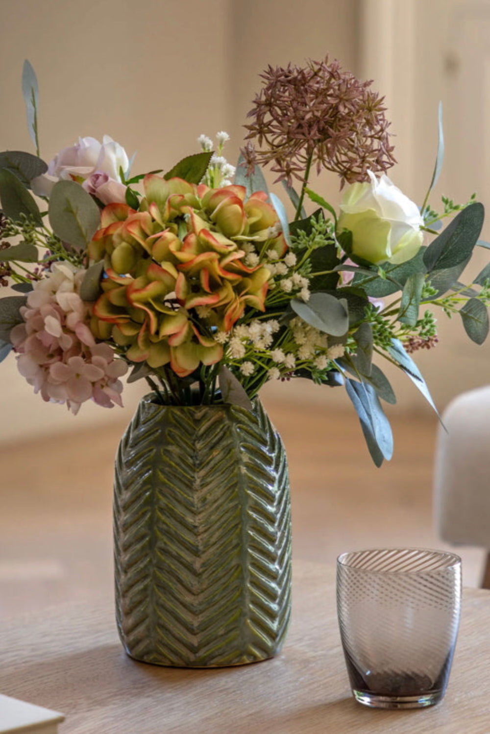 Bouquet of flowers in a green vase on a wooden table with a glass beside it.
