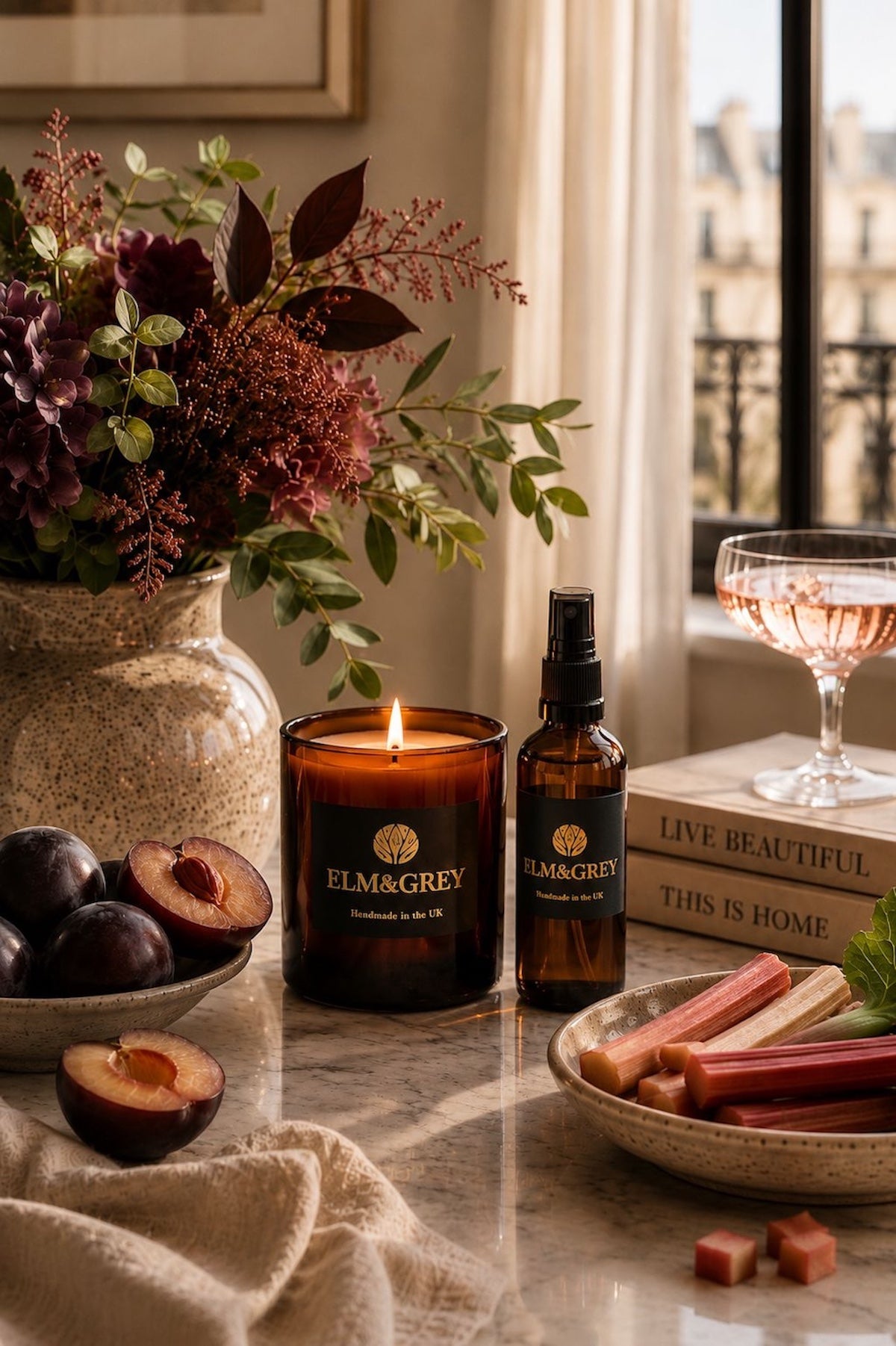 Elm & Grey Black Plum and Rhubarb scented candle and room spray on marble surface with dark purple florals, plums, rhubarb and rosé glass in Parisian setting