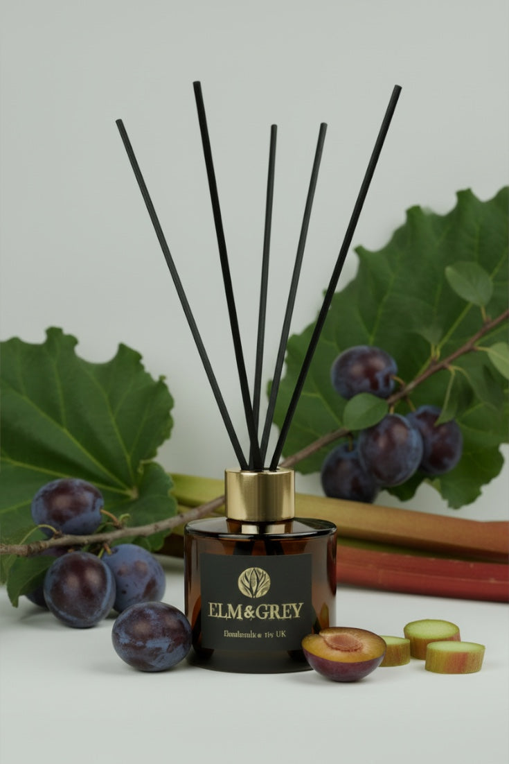 Reed diffuser with black sticks and gold cap on a white surface with plums and leaves.