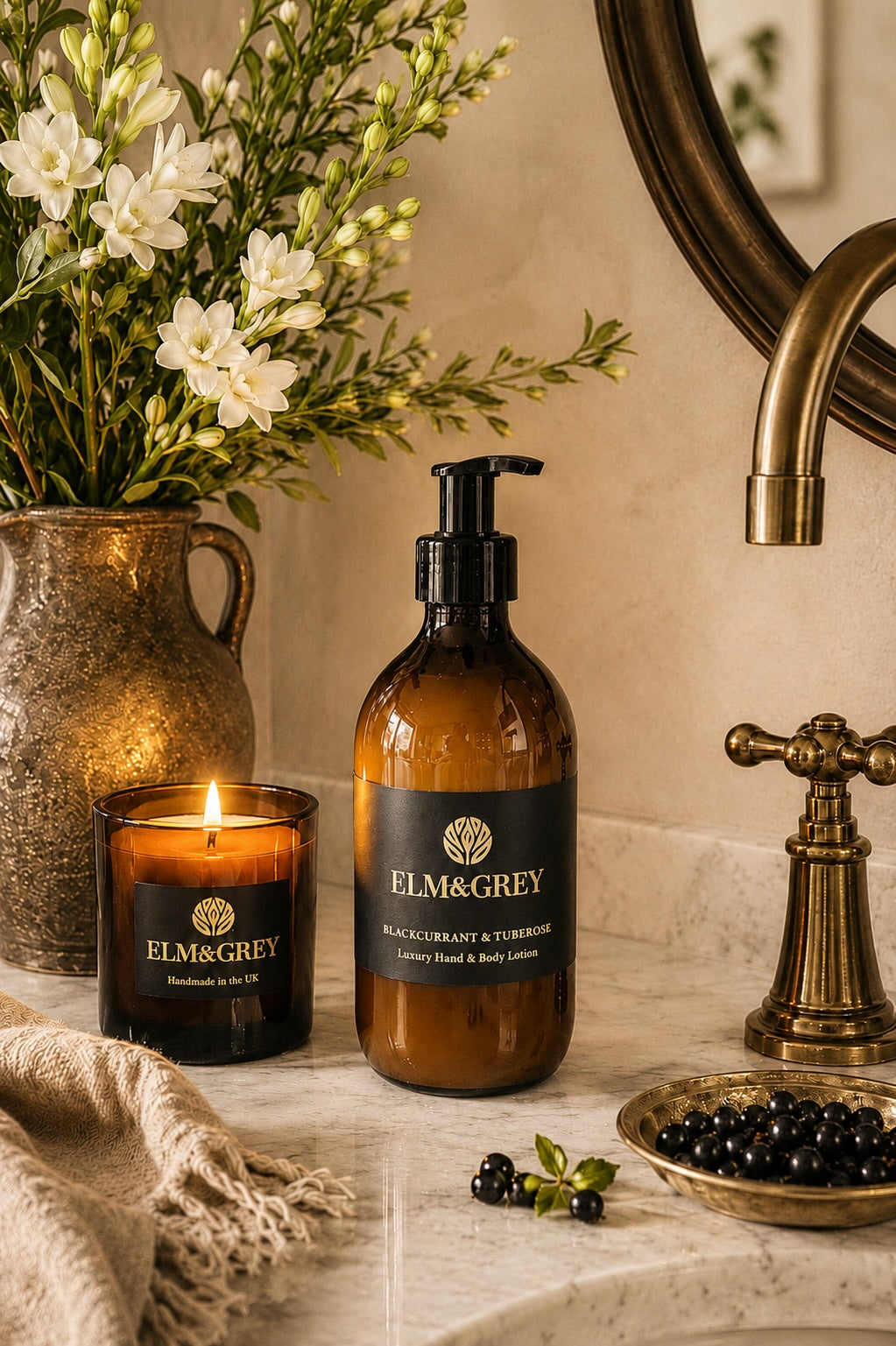 Elm & Grey Blackcurrant and Tuberose luxury hand and body lotion on marble bathroom surface with lit candle, white tuberose flowers and blackcurrants beside brass tap