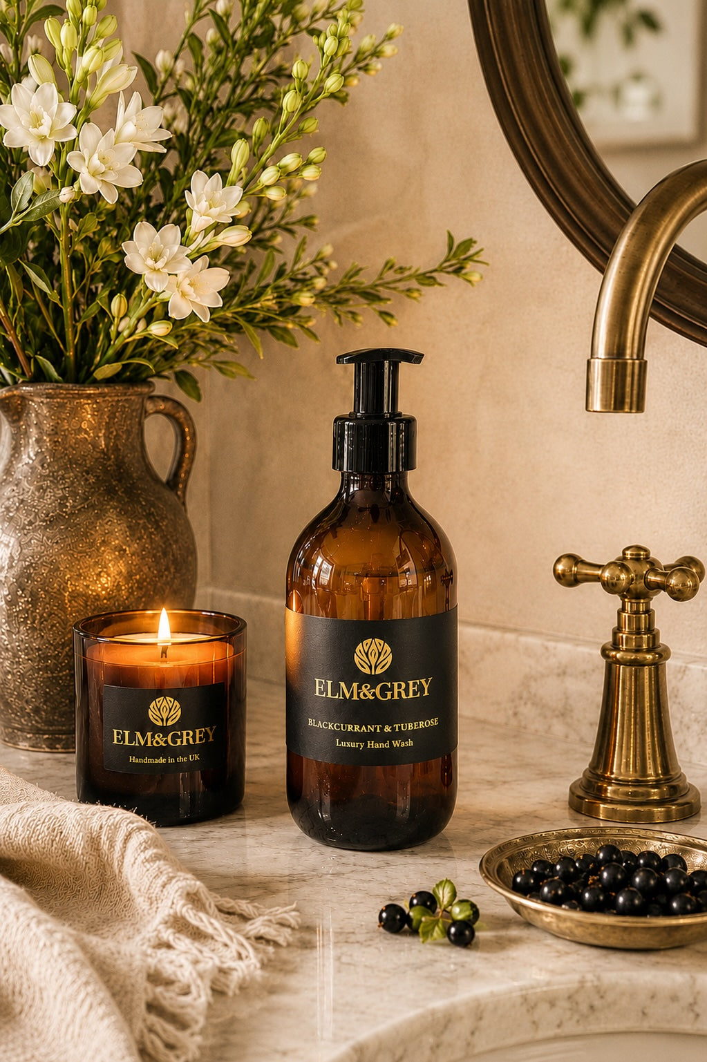 Elm & Grey Blackcurrant and Tuberose luxury hand wash on marble bathroom surface with lit candle, white tuberose flowers and blackcurrants beside brass tap in candlelight