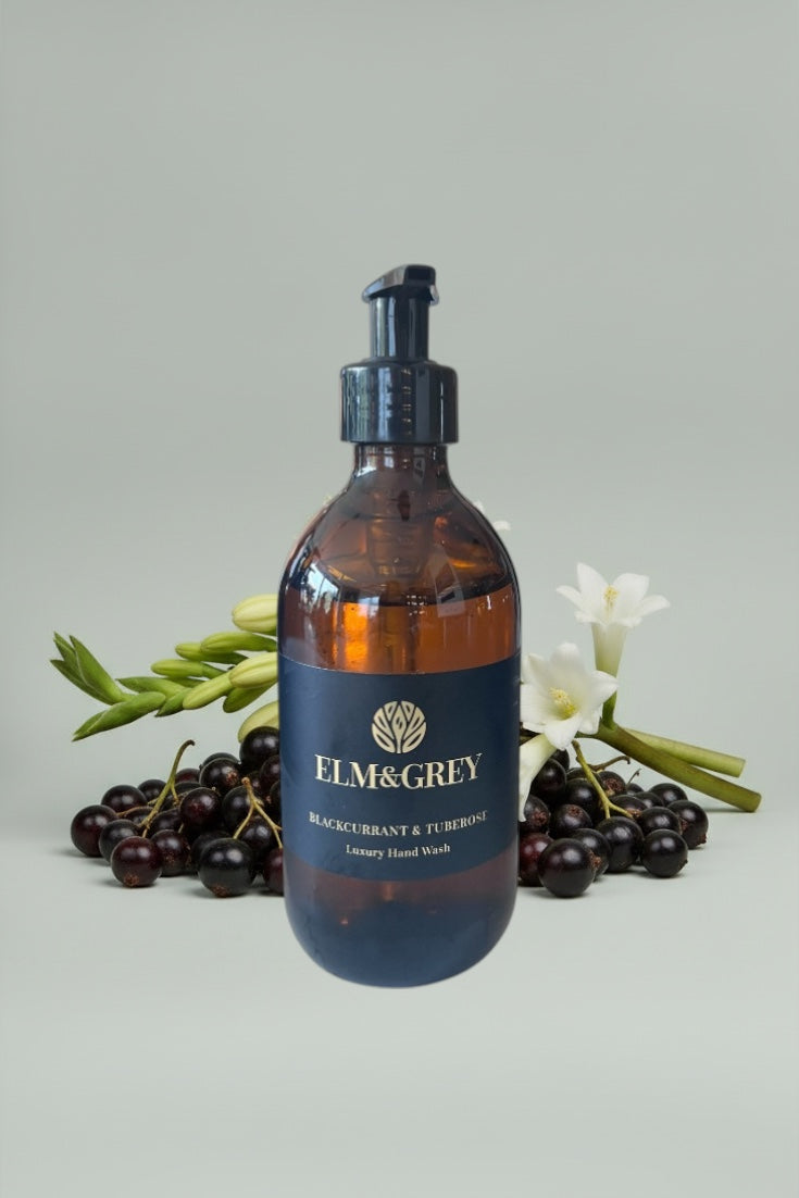 Gloss bottle of Elm & Grey blackcurrant and turmeric hand wash with blackcurrants and flowers on a light gray background