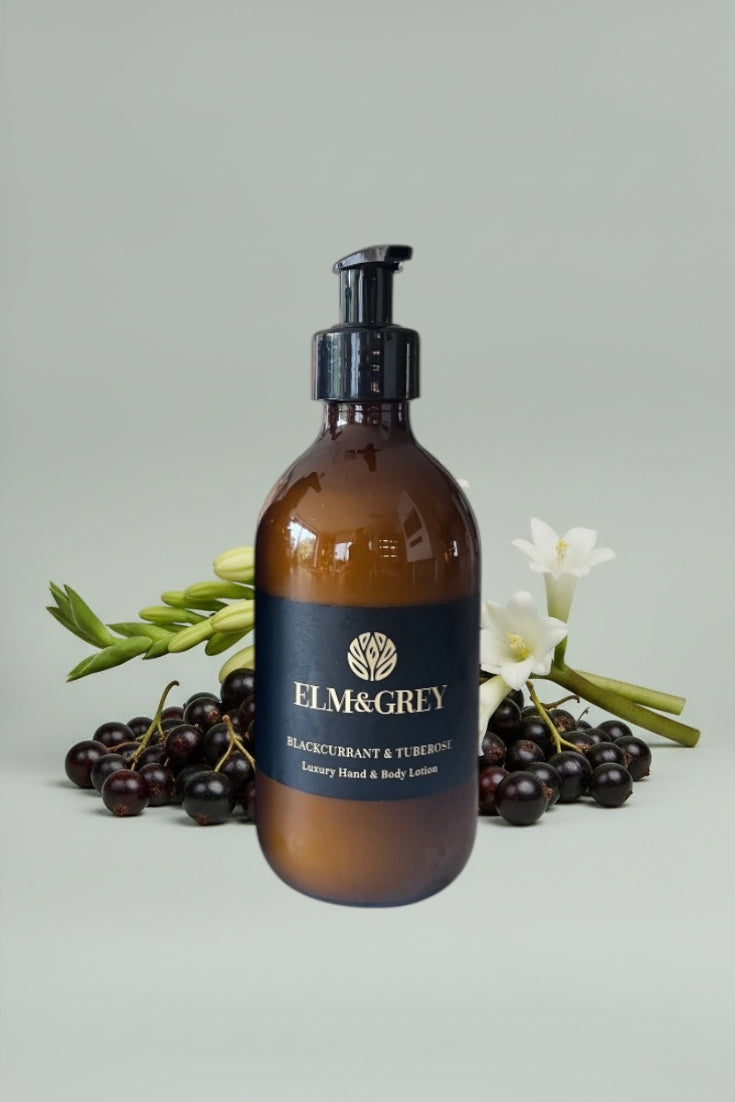 Amber bottle of Elm & Grey Blackcurrant & Tuberose lotion with blackcurrants and flowers on a light grey background