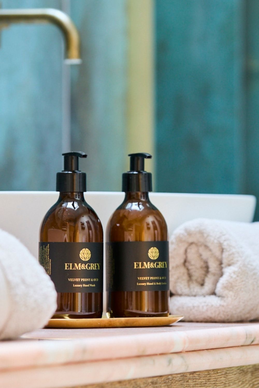Two brown bottles with black labels on a marble surface with towels and a sink in the background.