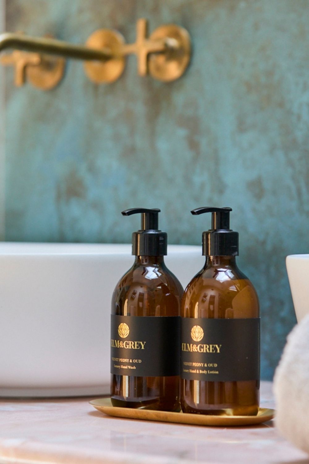 Two bottles of Elm & Grey soap and lotion on a tray with a bathroom setting in the background.