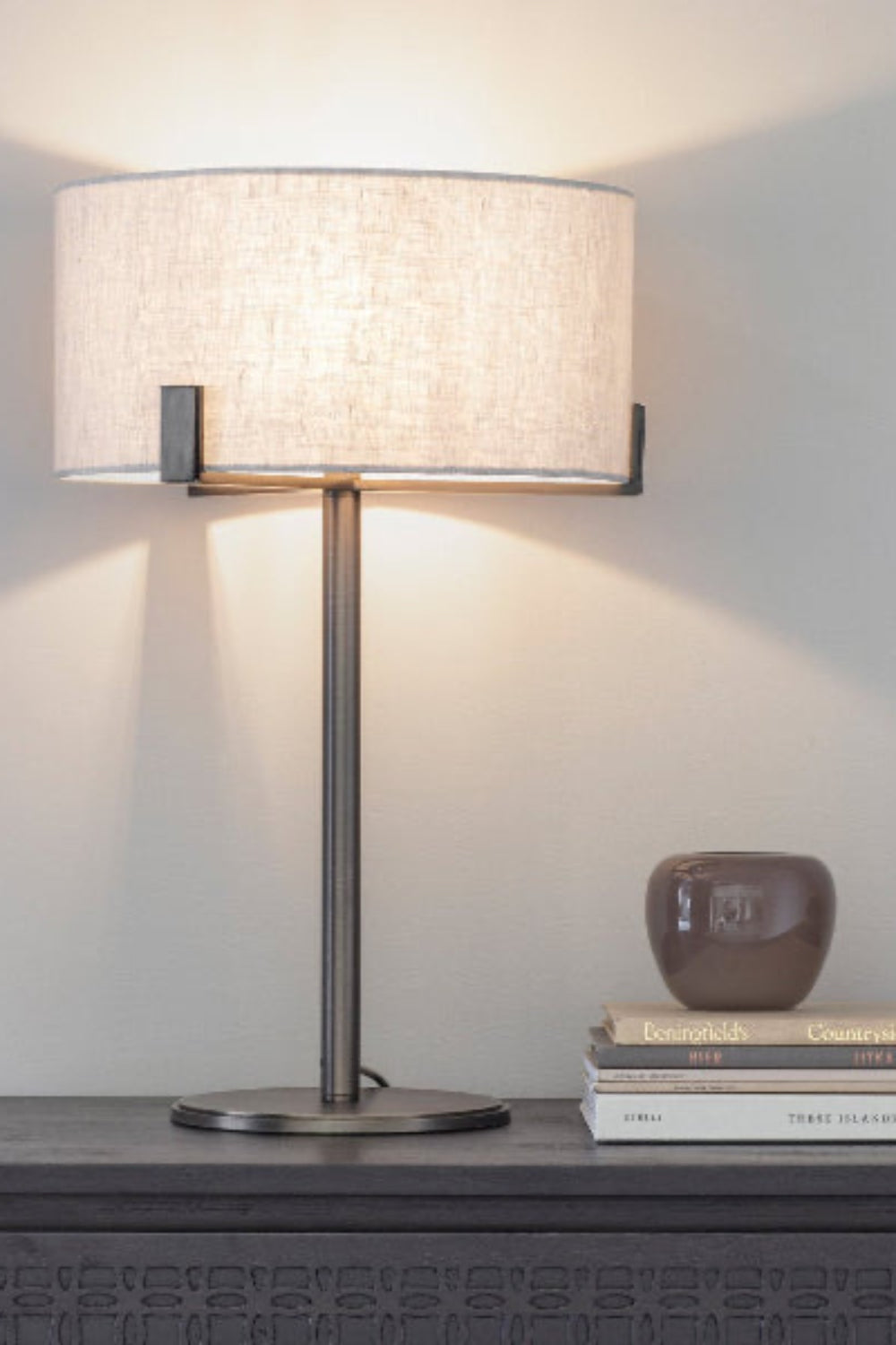 Brushed bornze table lamp with a natural linen textured shade on a surface with books and a small object. Elm & Grey.