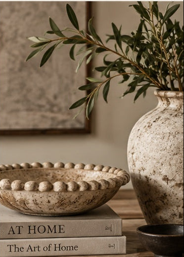 A styled home decor scene featuring a large speckled beige ceramic vase with green olive branches, a matching scalloped ceramic bowl resting on two books, and a small dark bowl in the foreground against a warm neutral wall.