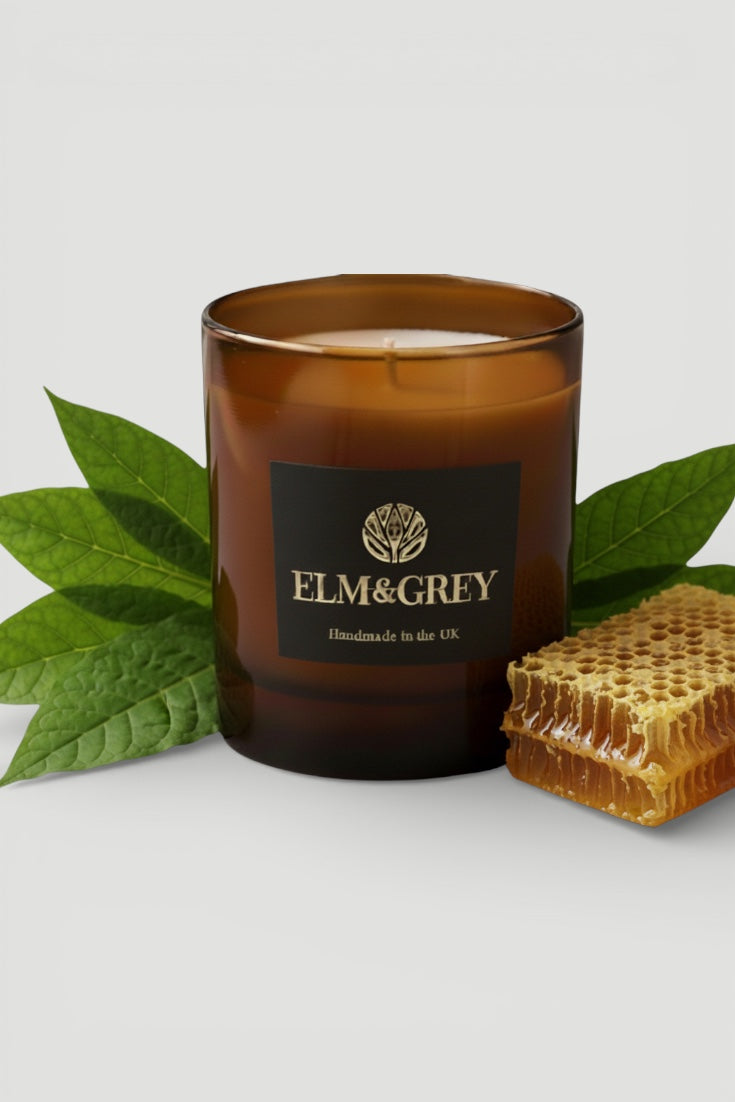dark honey and tobacco leaf candle with Elm & Grey branding, surrounded by green leaves and a honeycomb on a light gray background