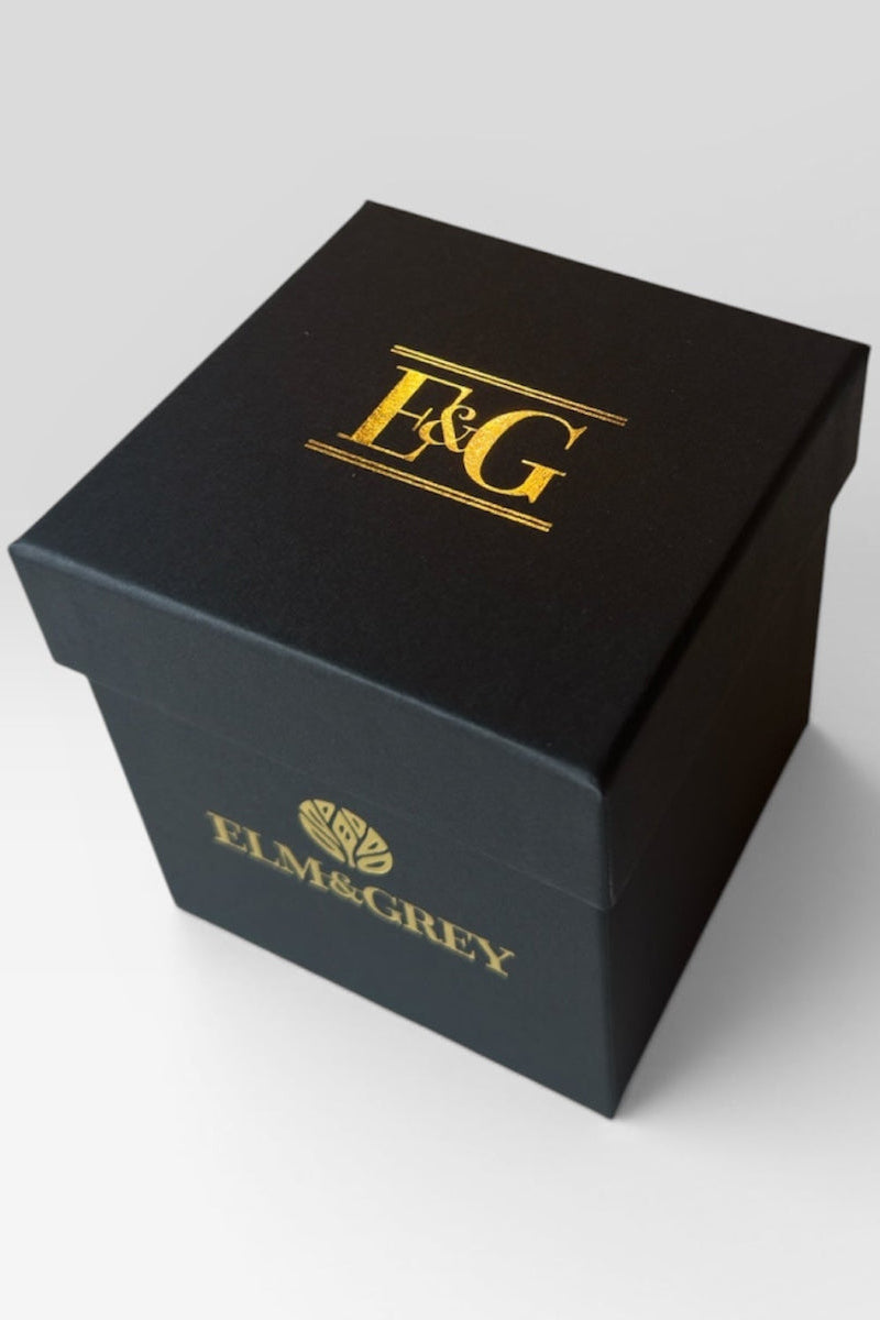 Elm & Grey matte black gift box with gold E&G monogram foil on lid and gold Elm & Grey logo on side, product shot on grey background