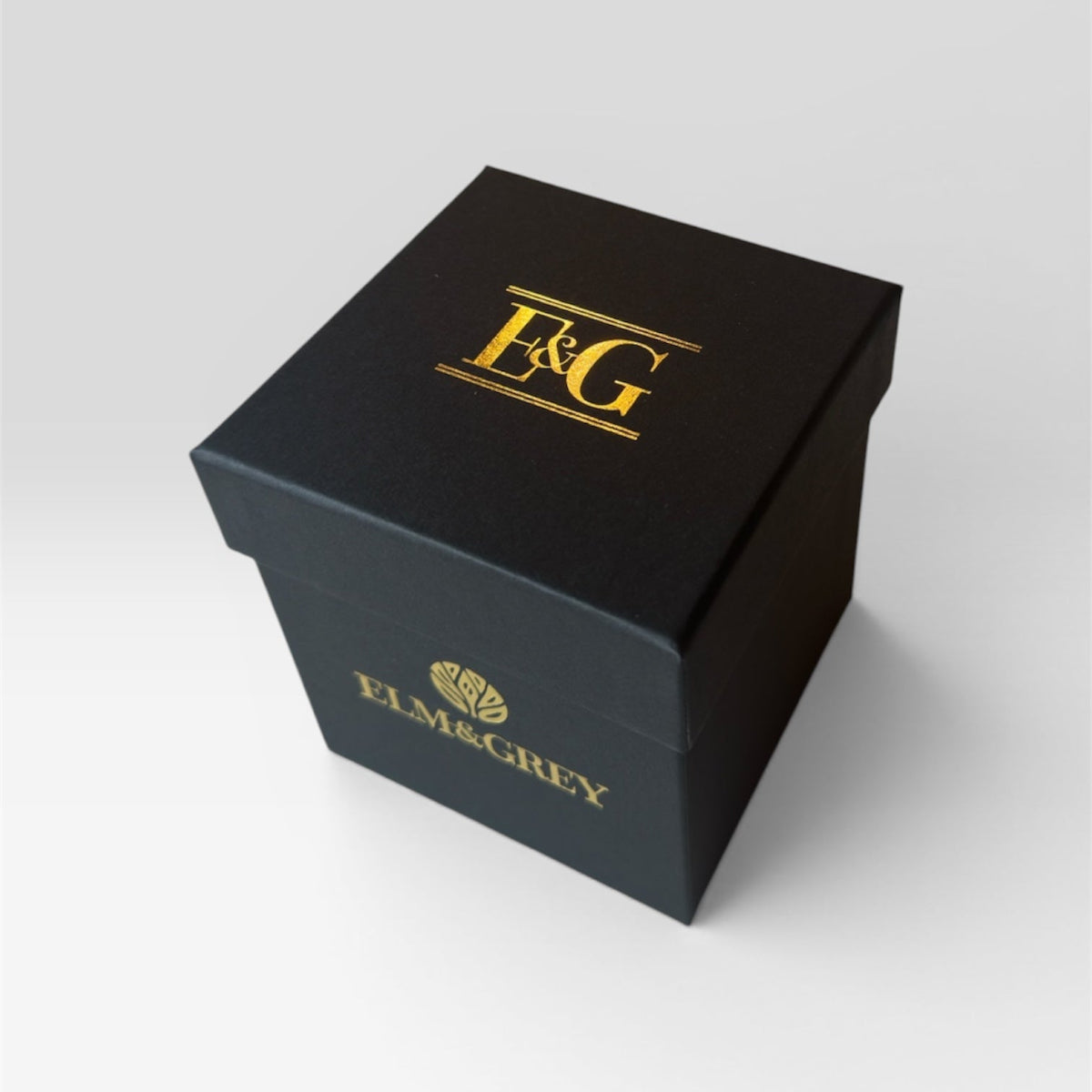 Elm & Grey matte black gift box with gold E&G monogram foil on lid and gold Elm & Grey logo on side, product shot on grey background
