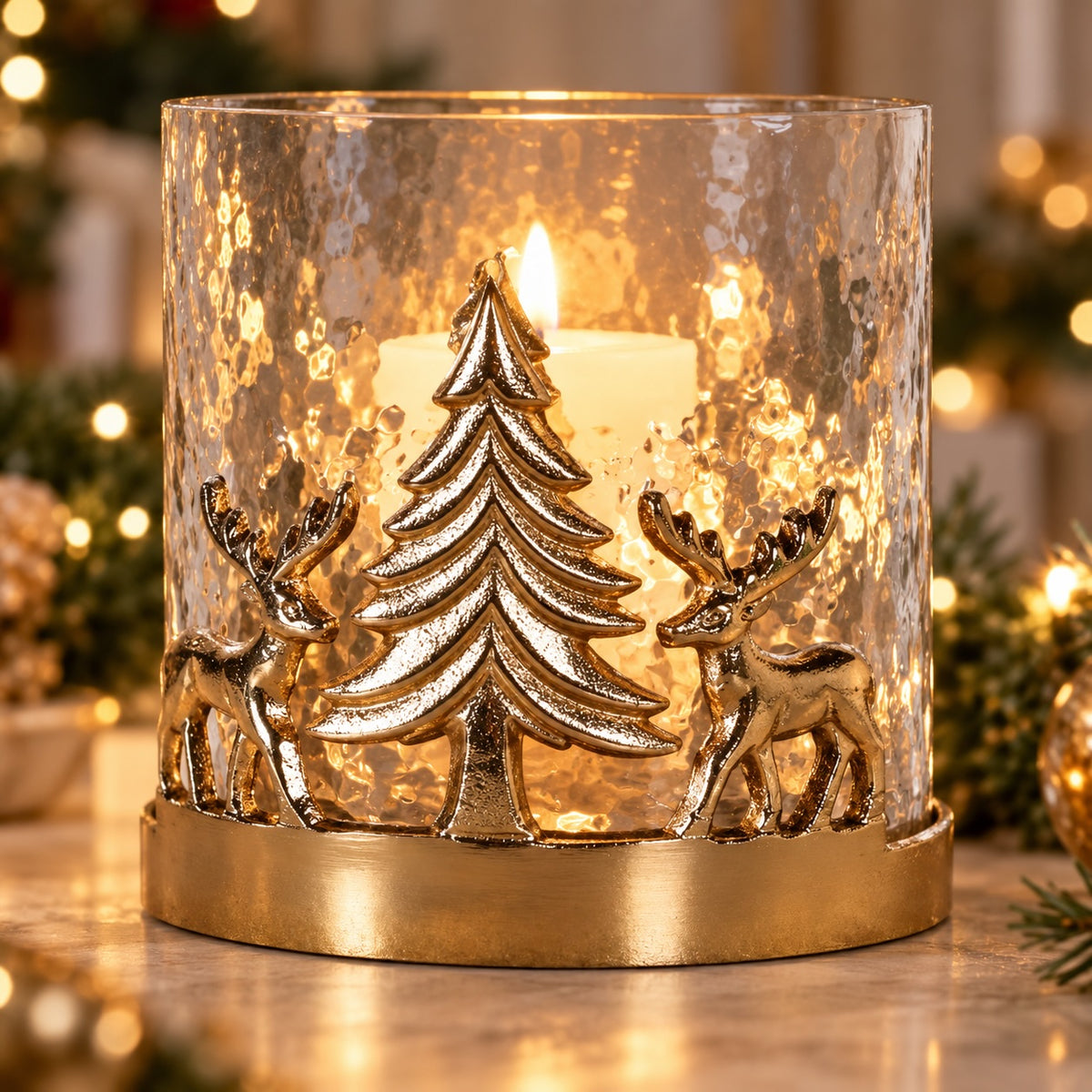 A festive holiday candle holder with a hammered clear glass cylinder, featuring a gold metal base with a Christmas tree and two deer silhouette design, lit with a white candle inside.