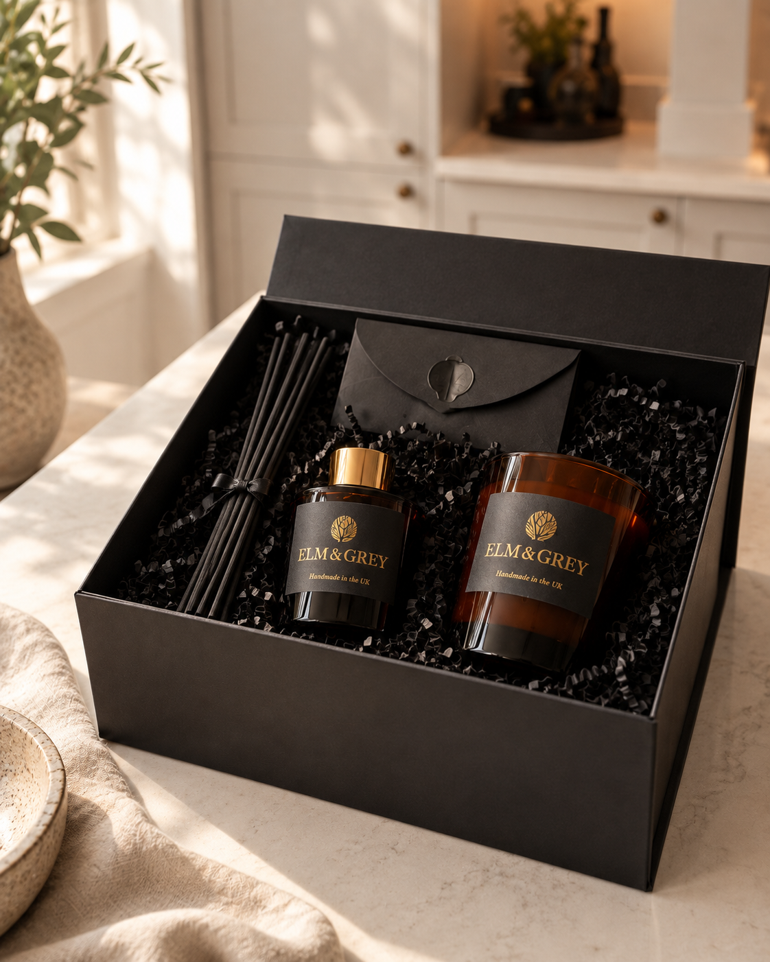 An open black gift box on a light countertop containing a black reed diffuser bottle with a gold cap, a brown candle jar with a black label, black reeds, and a black envelope. The box is branded ELM & GREY.