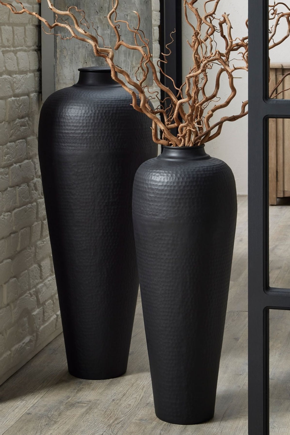 Two black hammered textured vases with dried twisted willow branches against a brick wall.