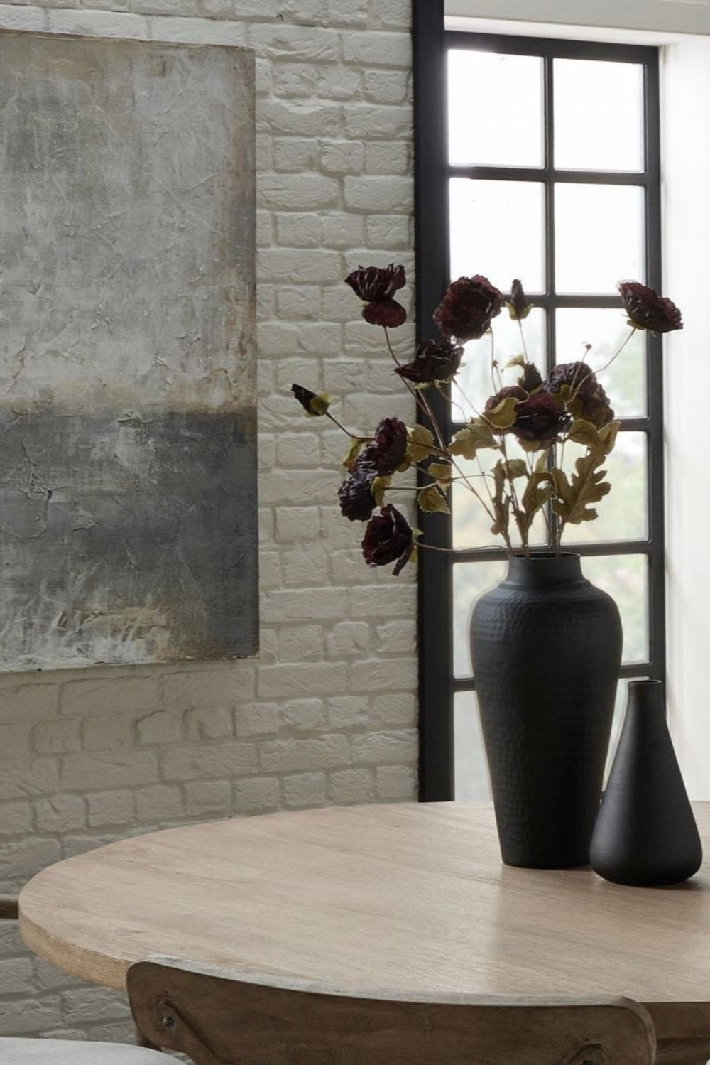 Matt black hammered vase with dried flowers on a wooden table against a brick wall.