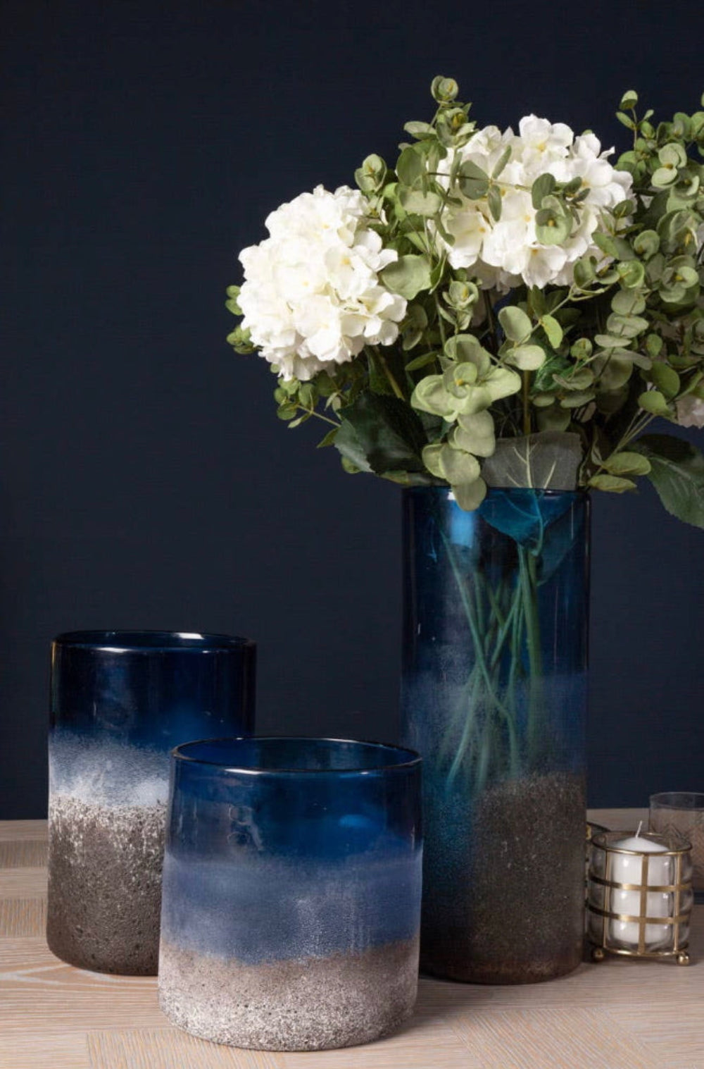 Three blue sand effect textured gradient glass vases with white and green flowers on a dark background.