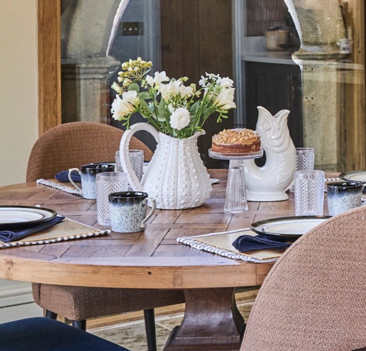 A kitchen table set with a cake on a glass stand a white urchin jug with flowers in it and a gluggle jug in white