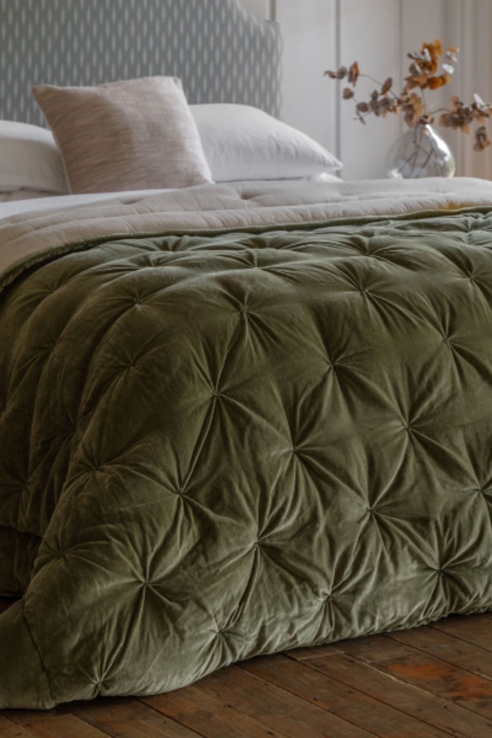 Green textured comforter on a bed with pillows and a vase of flowers in the background.