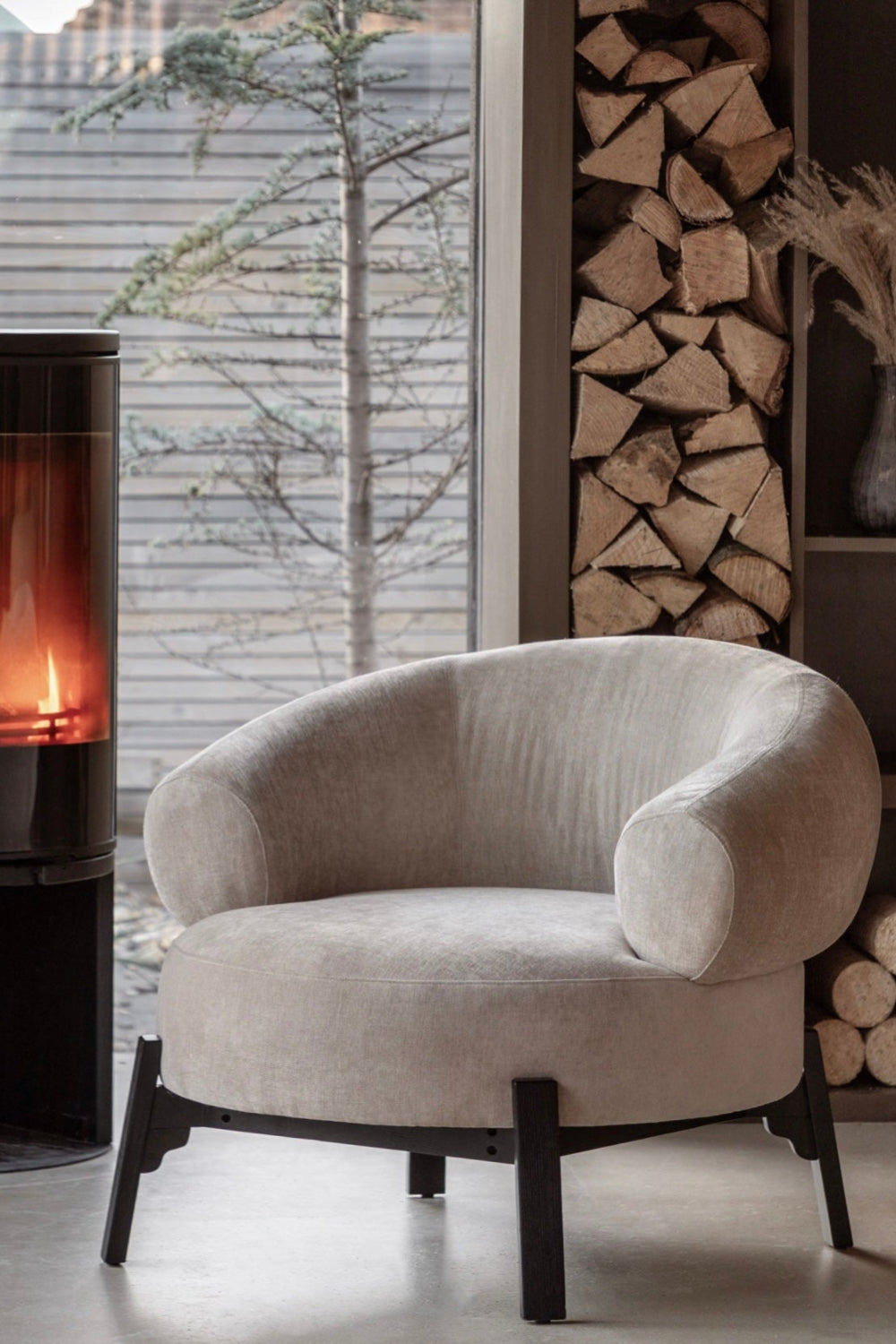 Modern curved armchair in a room with a stove and stacked firewood, Elm & Grey.