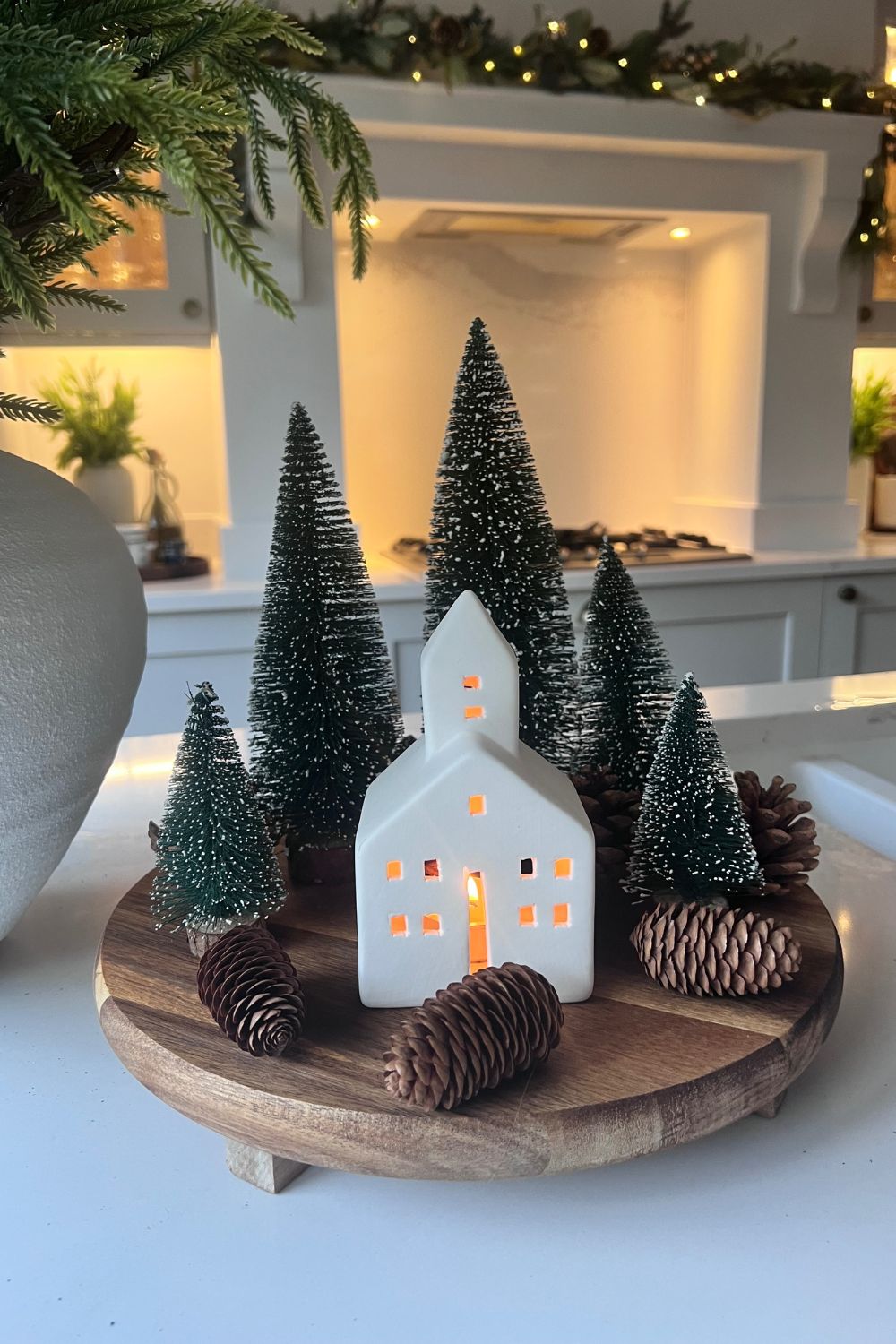 Decorative setup with a white ceramic house, pine trees, and pinecones on a wooden stand in a kitchen.