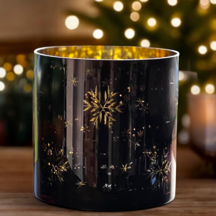 Navy candle holder with gold snowflake design in front of a Christmas tree with lights.