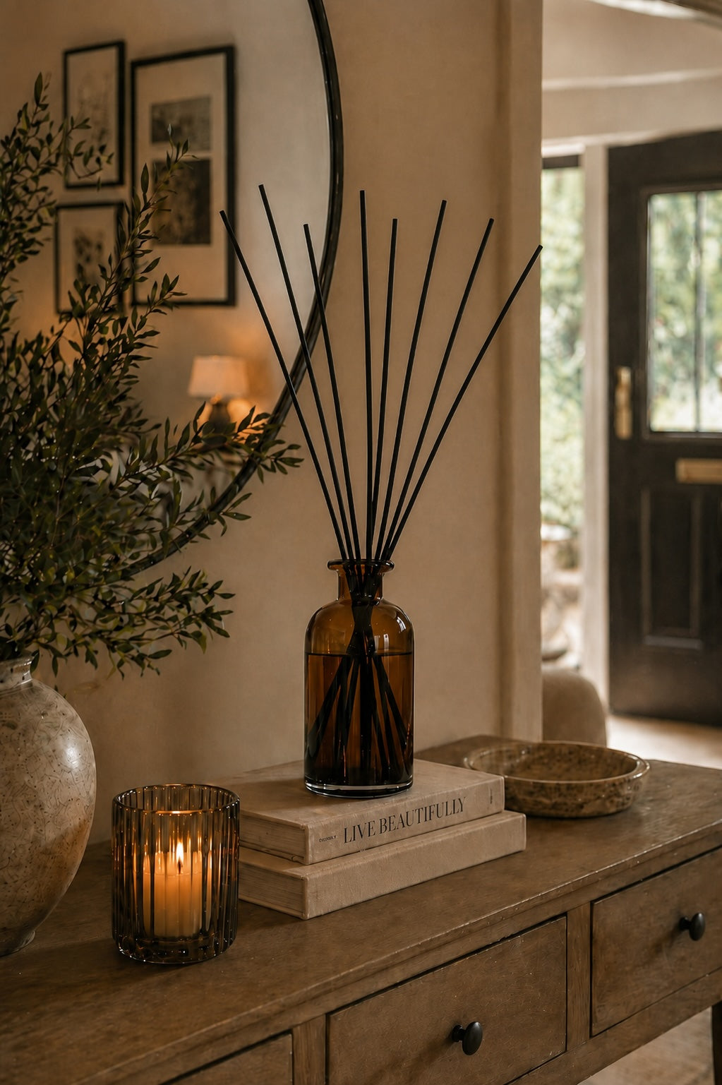 An amber glass reed diffuser bottle with black reeds sits on stacked books on a wooden console table in a warm, styled interior. A lit candle, a large vase with greenery, and framed wall art are nearby.