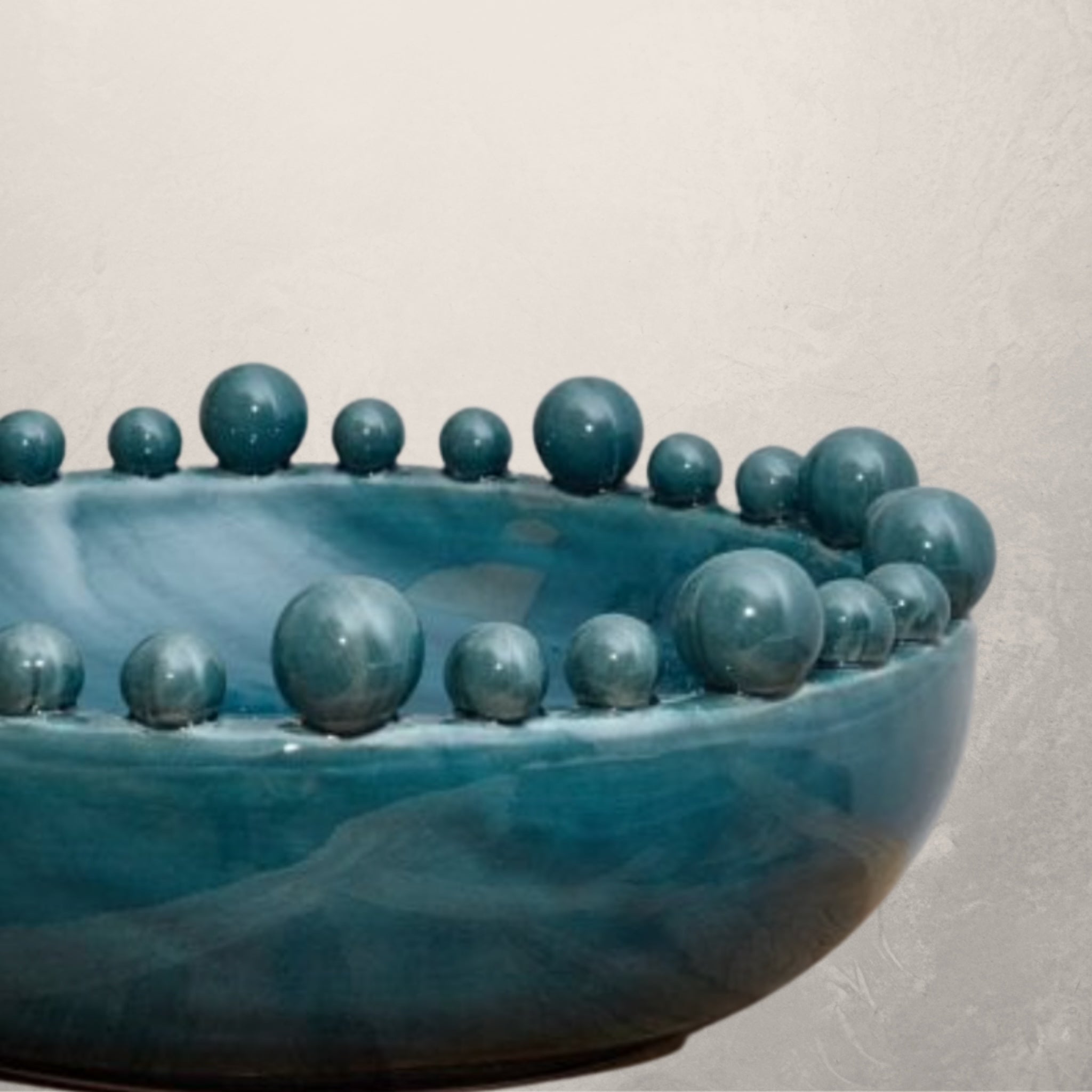A large handmade ceramic bowl with a teal color and bobble decorations on the rim, on a white background.. Elm & Grey.