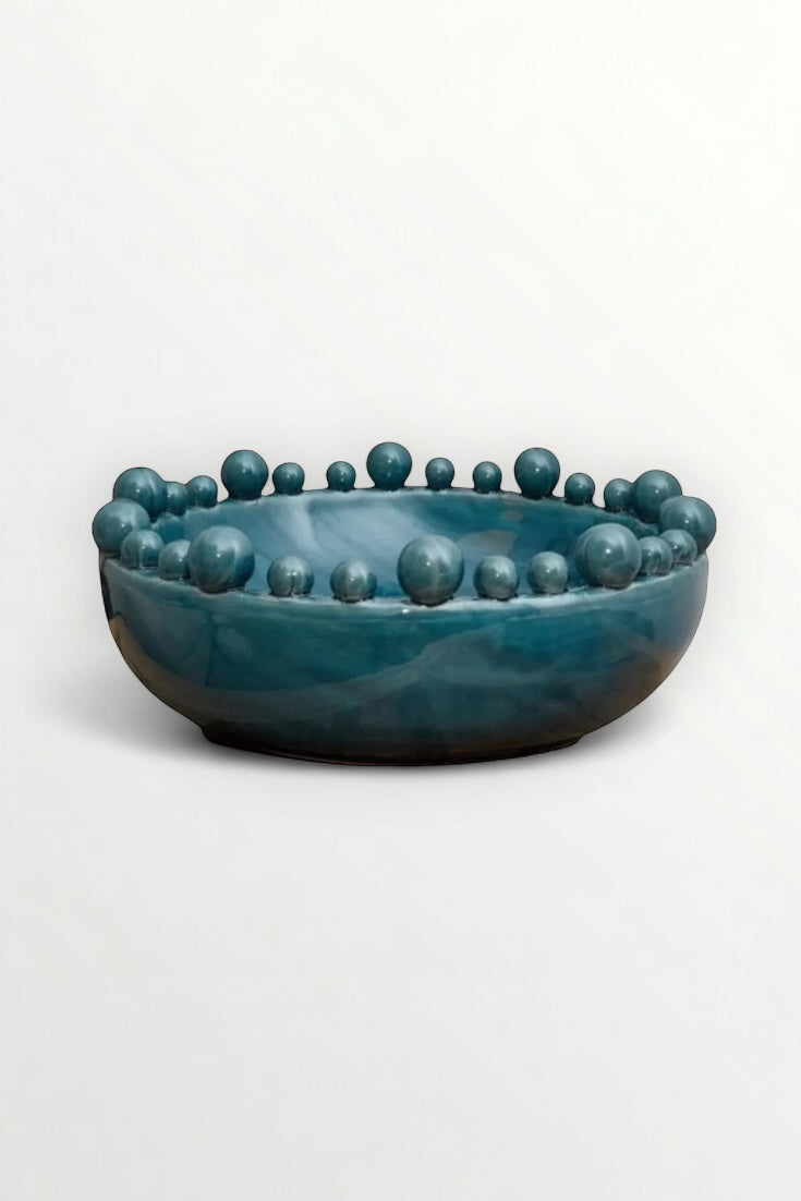 A large handmade ceramic bowl with a teal color and bobble decorations on the rim, on a white background.. Elm & Grey.
