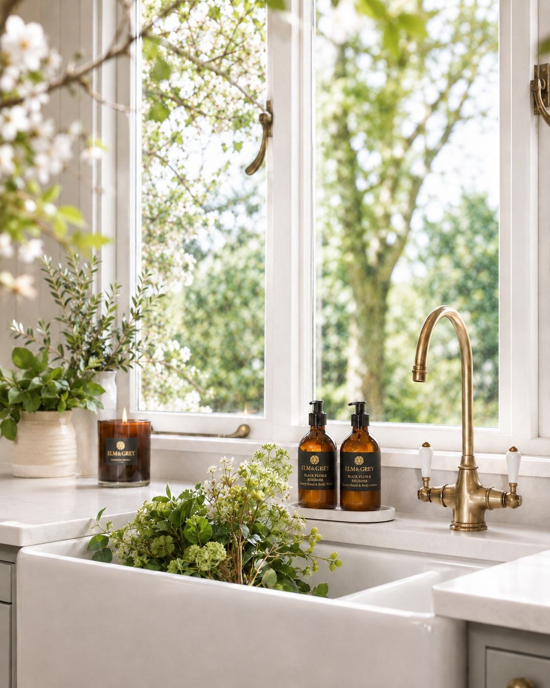 Two amber glass pump bottles with black labels and gold lettering sit on a white kitchen counter beside a brass faucet and a large white farmhouse sink. A window behind them shows green trees outside, and potted plants decorate the counter.