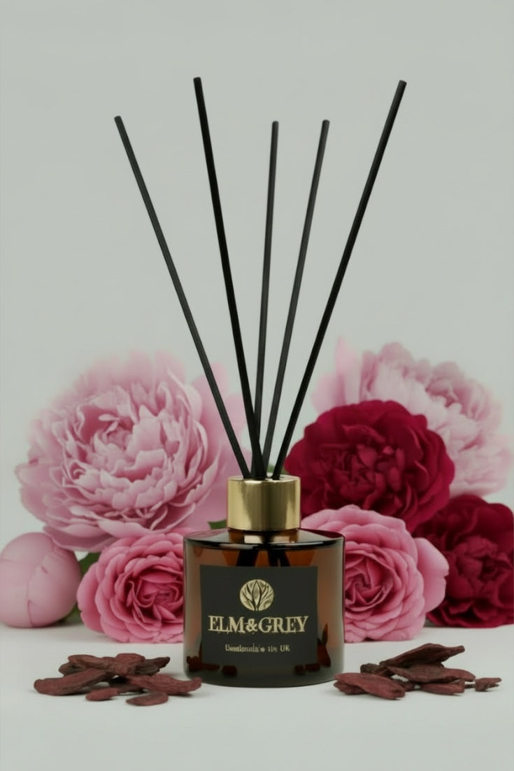 Reed diffuser with black sticks in a decorative container surrounded by pink and red flowers on a light gray background.