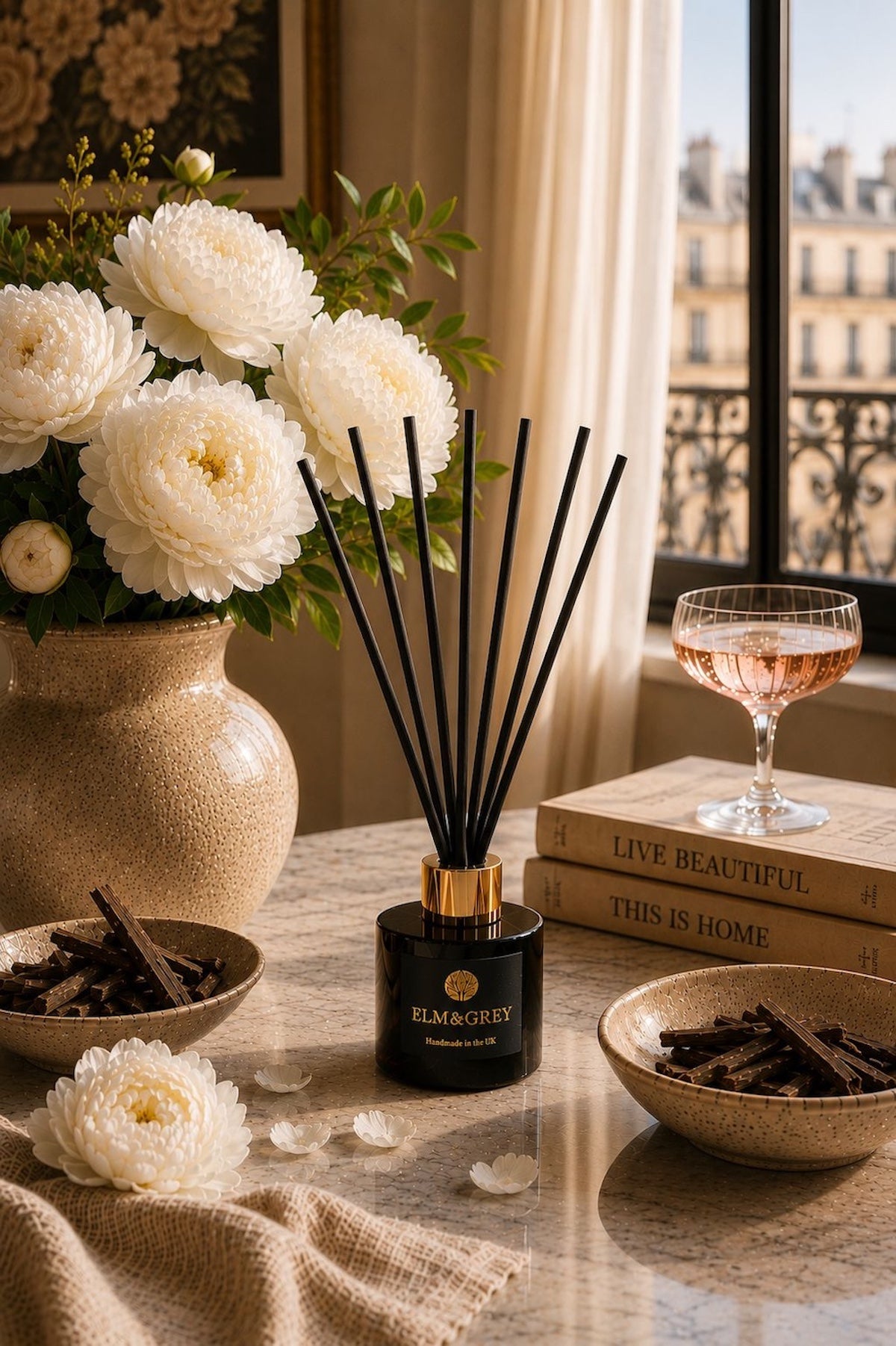 Elm & Grey Velvet Peony and Oud reed diffuser on marble surface with white peonies, oud wood chips and a Parisian window view