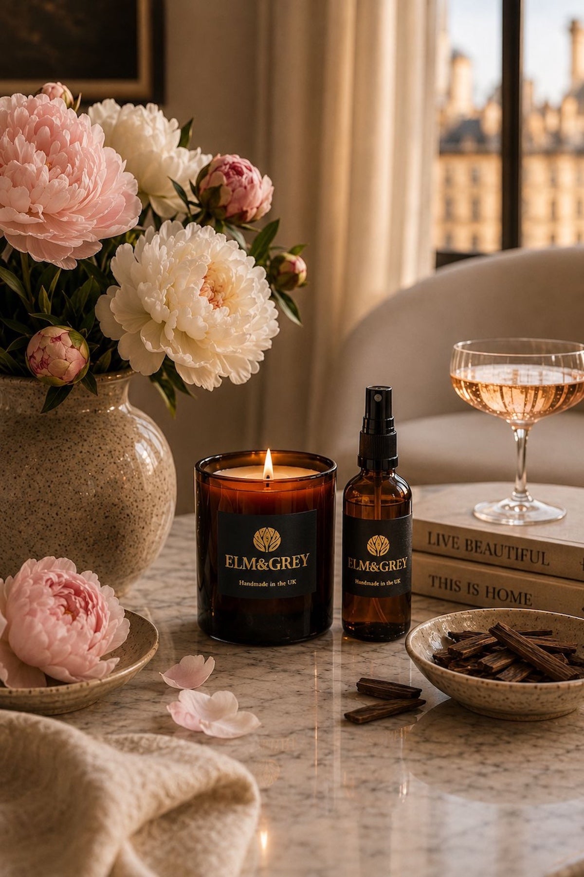 Elm & Grey Velvet Peony and Oud scented candle and room spray on a marble table with pink peonies, oud wood chips and a glass of rosé in a Parisian setting