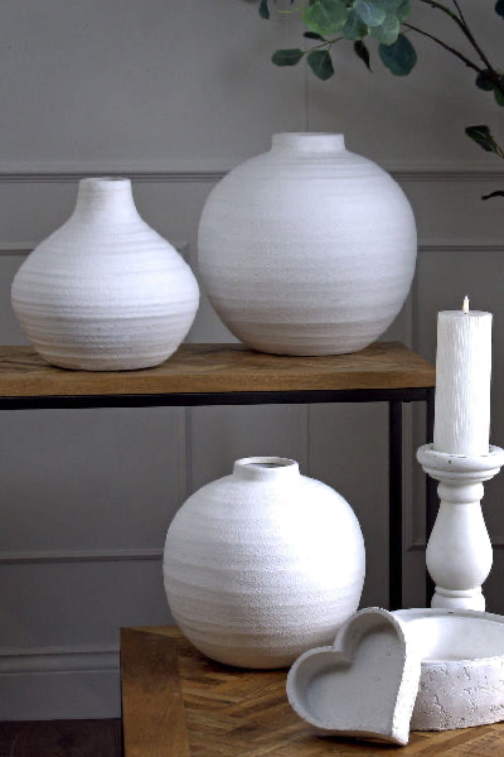 Three white ceramic vases on a wooden surface with a candle and heart-shaped object in the foreground.