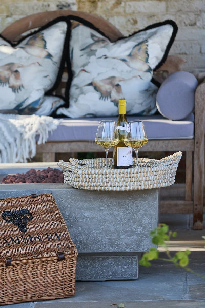 Outdoor setting with a bottle of wine and glasses on stacked seagrass trays on a stone table with cushions and a basket in the background., Elm & Grey.