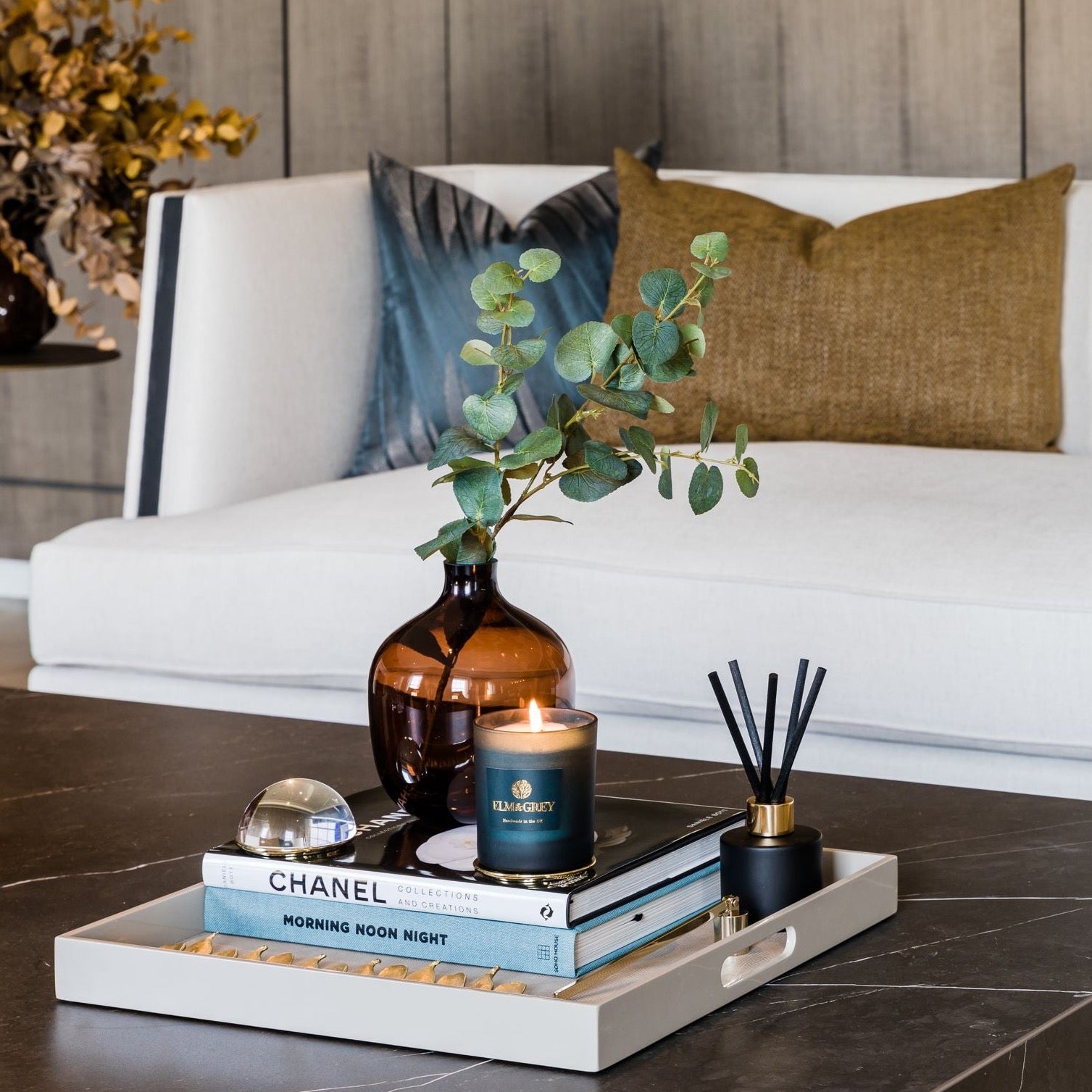 A pre-styled lacquered tray with accessories including a vase with a plant, coffee table books, a candle, and a diffuser, placed on a coffee table in a room setting.