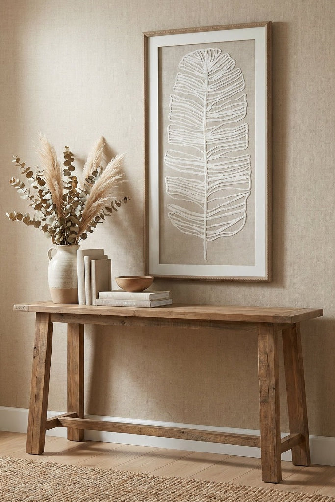 A framed wall art print featuring a white feather design on a beige background, styled on a rustic wooden console table with books, a small bowl, and a vase of dried grasses against a textured beige wall.