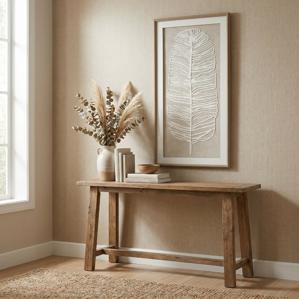 Wooden console table with decorative items against a beige wall with a framed abstract leaf art piece.