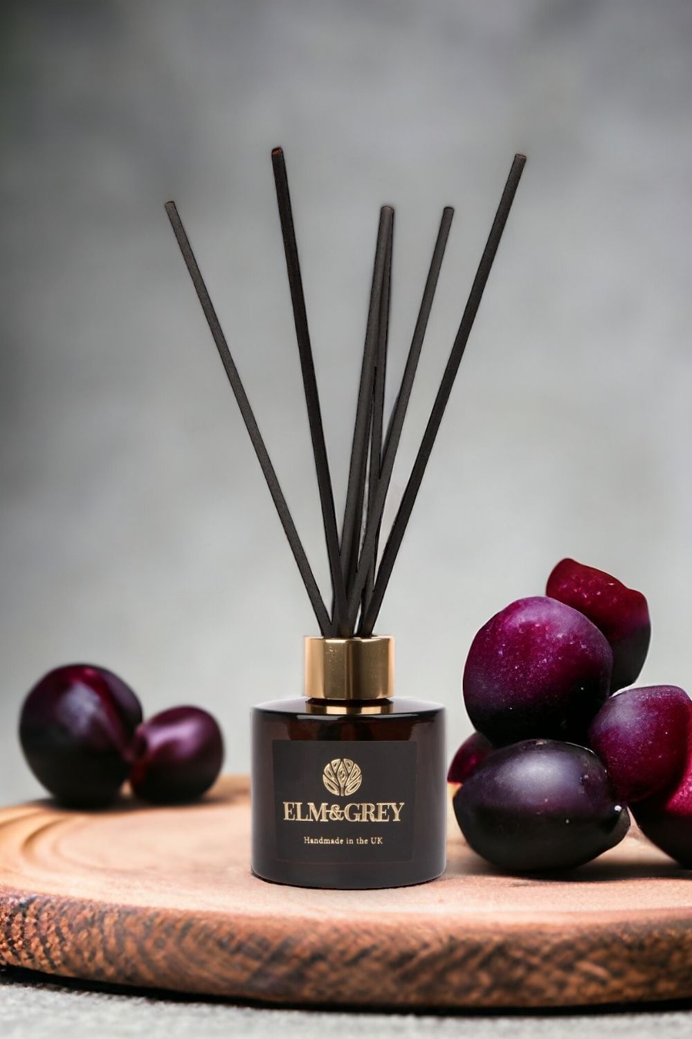 Reed diffuser with black sticks and a dark container on a wooden board with fruits, branded 'Elm & Grey'.